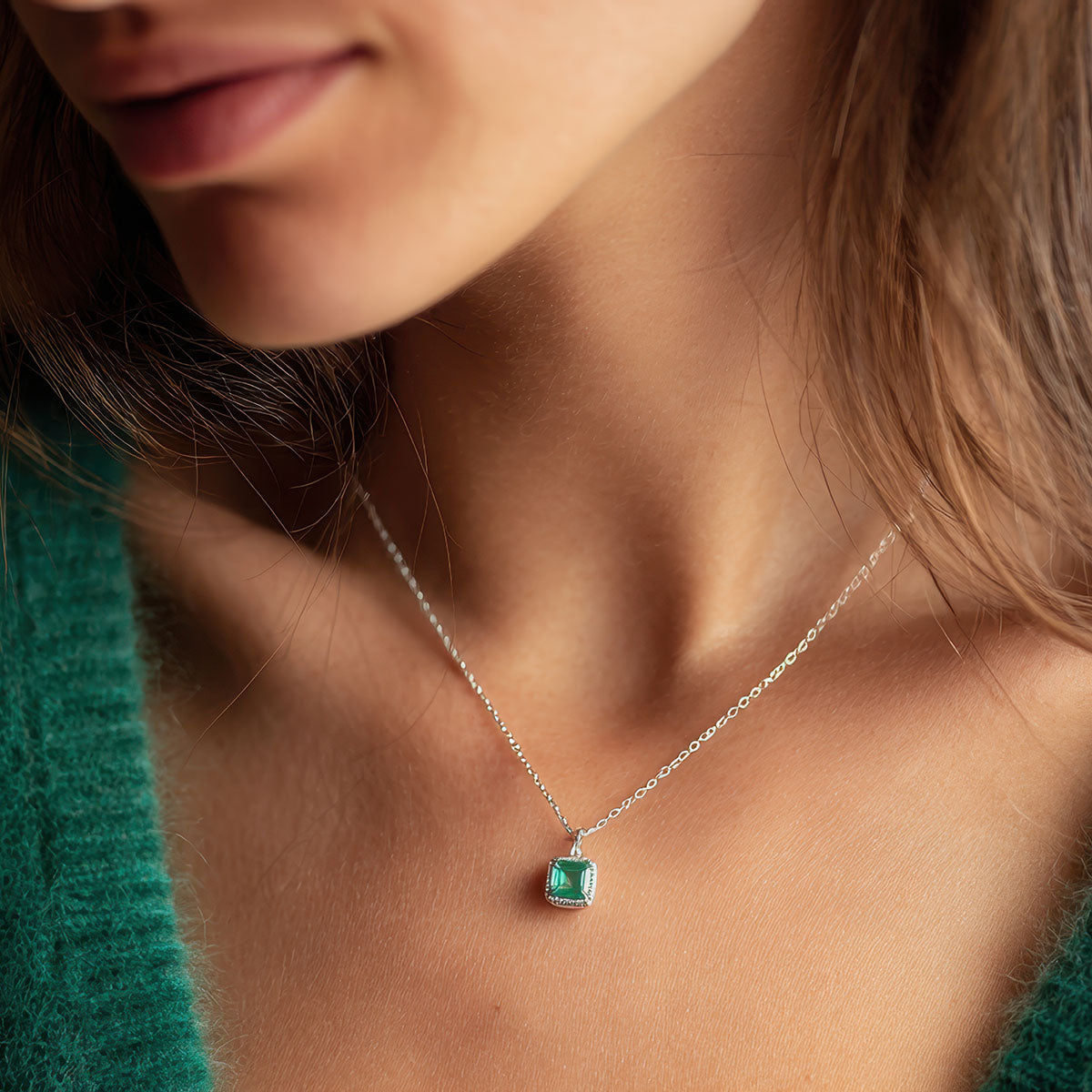 Sieviete ar smalku sudraba krāsas kaklarotu un kvadrātveida kulonu, kurā iestrādāts zaļš akmens.