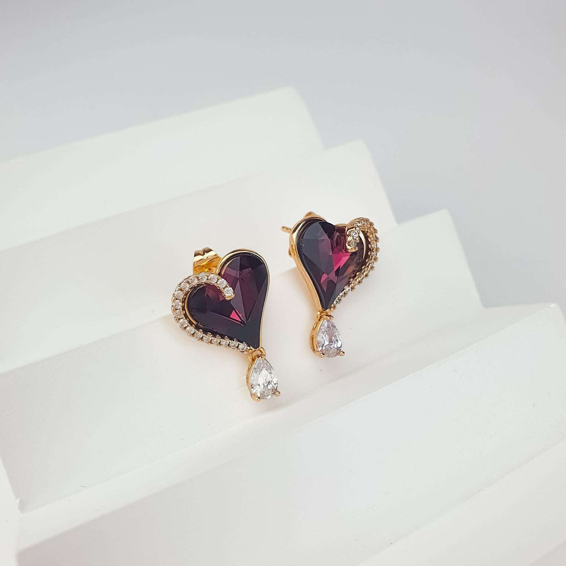 Серьги-сердца с фиолетовым кристаллом под углом на белом подиуме с видимой застёжкой-гвоздиком